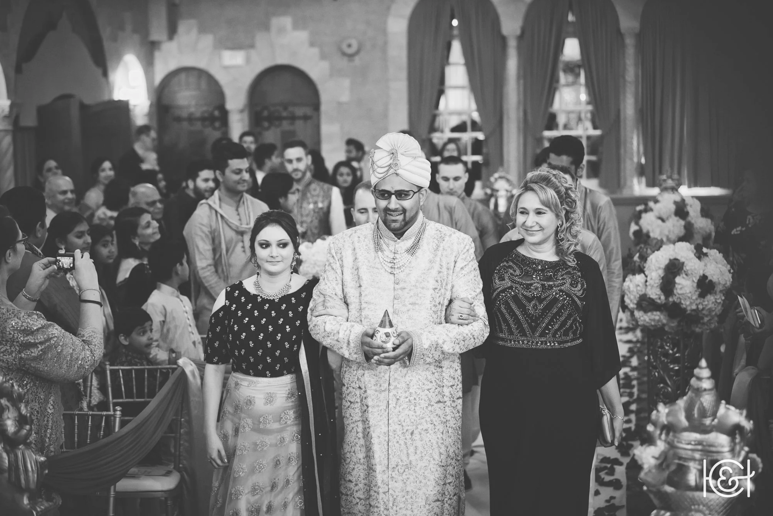 A traditional Indian wedding ceremony in a decorated hall with people dressed in ethnic attire. The groom, wearing a sherwani and turban, stands with a smile, holding a ceremonial object. Two women stand with him, one on each side, all smiling. Guest