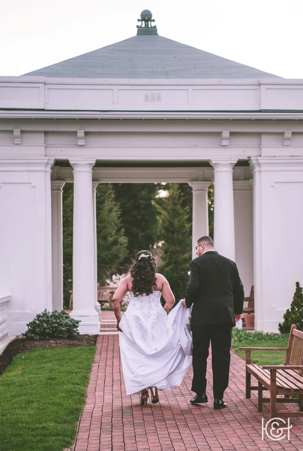 A bride and groom walk together on a brick pathway through a white pavilion with columns, holding hands, with greenery in the background.