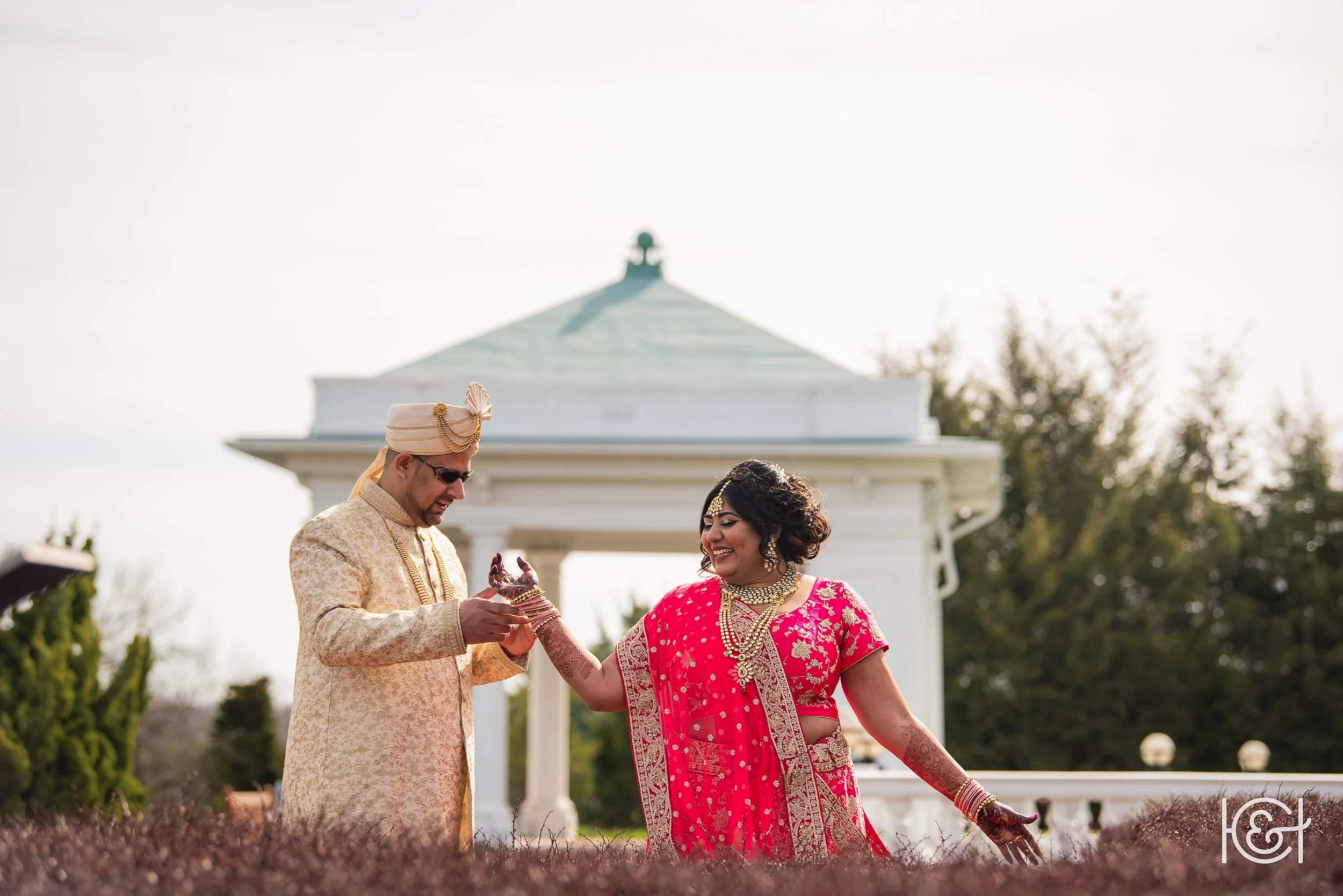 A South Asian bride and a groom in traditional wedding attire during a wedding ceremony outdoors, standing in front of a white pavilion with trees in the background, exchanging rings and smiling.