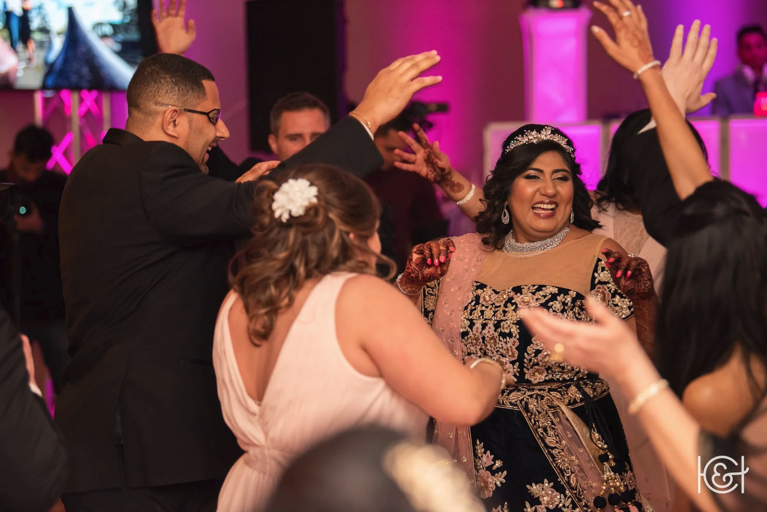 Group of people dancing and celebrating at a wedding reception, with a woman in traditional Indian attire and henna on her hands smiling and raising her hands.