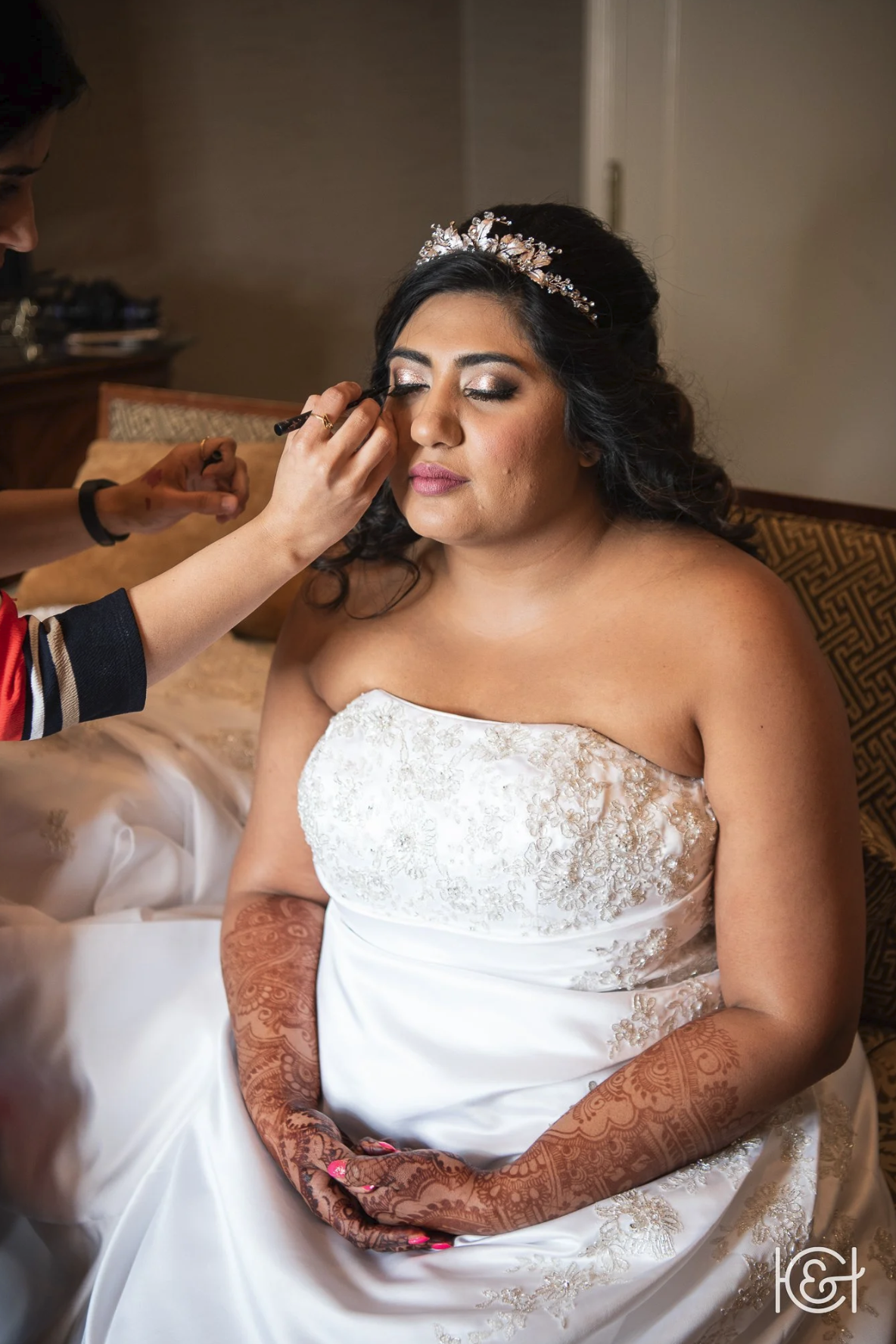 A bride is having makeup applied by a makeup artist. She is wearing a strapless white wedding gown with gold embroidery and a tiara, seated with her eyes closed, showing intricate henna designs on her hands and arms.