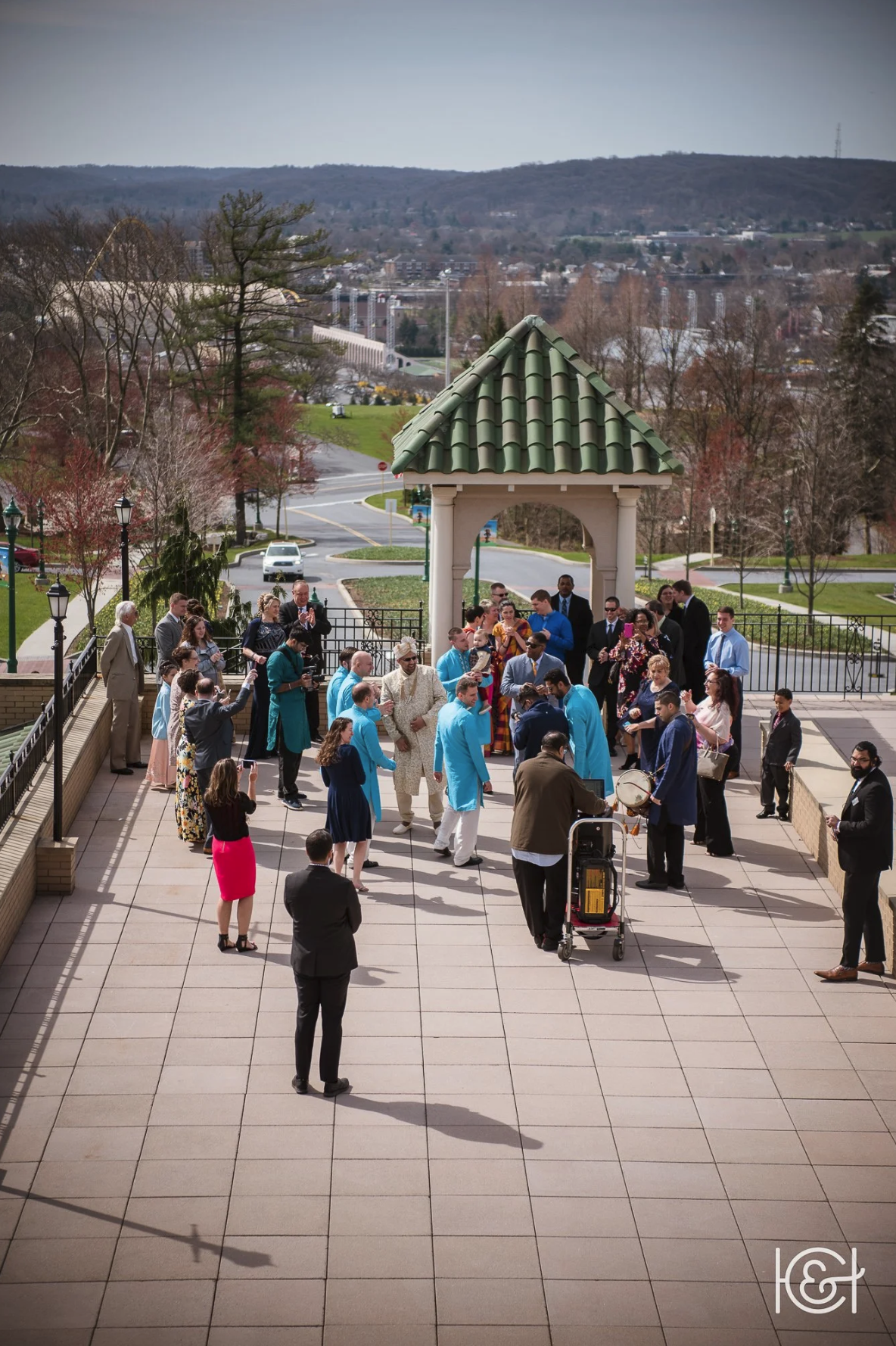 People gathered on a terrace outdoors, dressed in colorful formal attire, celebrating a wedding with some dancing and music, with a scenic view of rolling hills and a small town in the background.