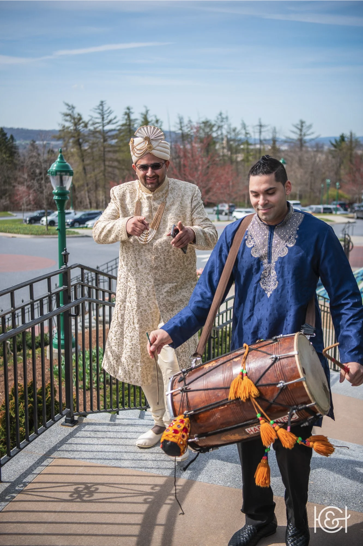 A man wearing traditional Indian attire, including a cream-colored sherwani and a turban, stands on a porch. He is smiling and holding a mobile phone. Another man dressed in a blue traditional outfit with embroidery is playing a large drum, decorated