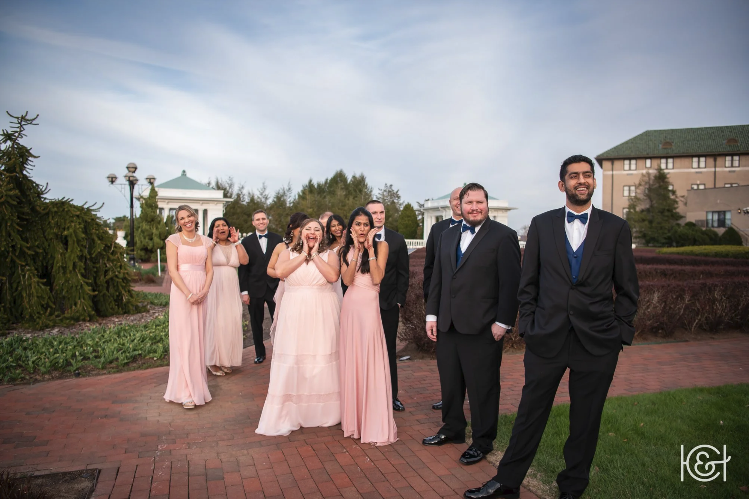 A group of people dressed in formal attire, including pink dresses and black tuxedos, standing outdoors on a brick path with trees, buildings, and a cloudy sky in the background, smiling and posing for a photo.