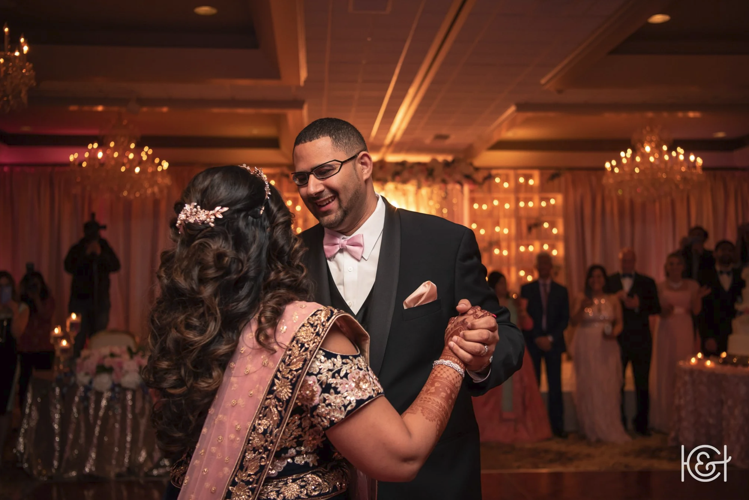 A couple dancing at a wedding reception, with guests in the background and warm lighting.