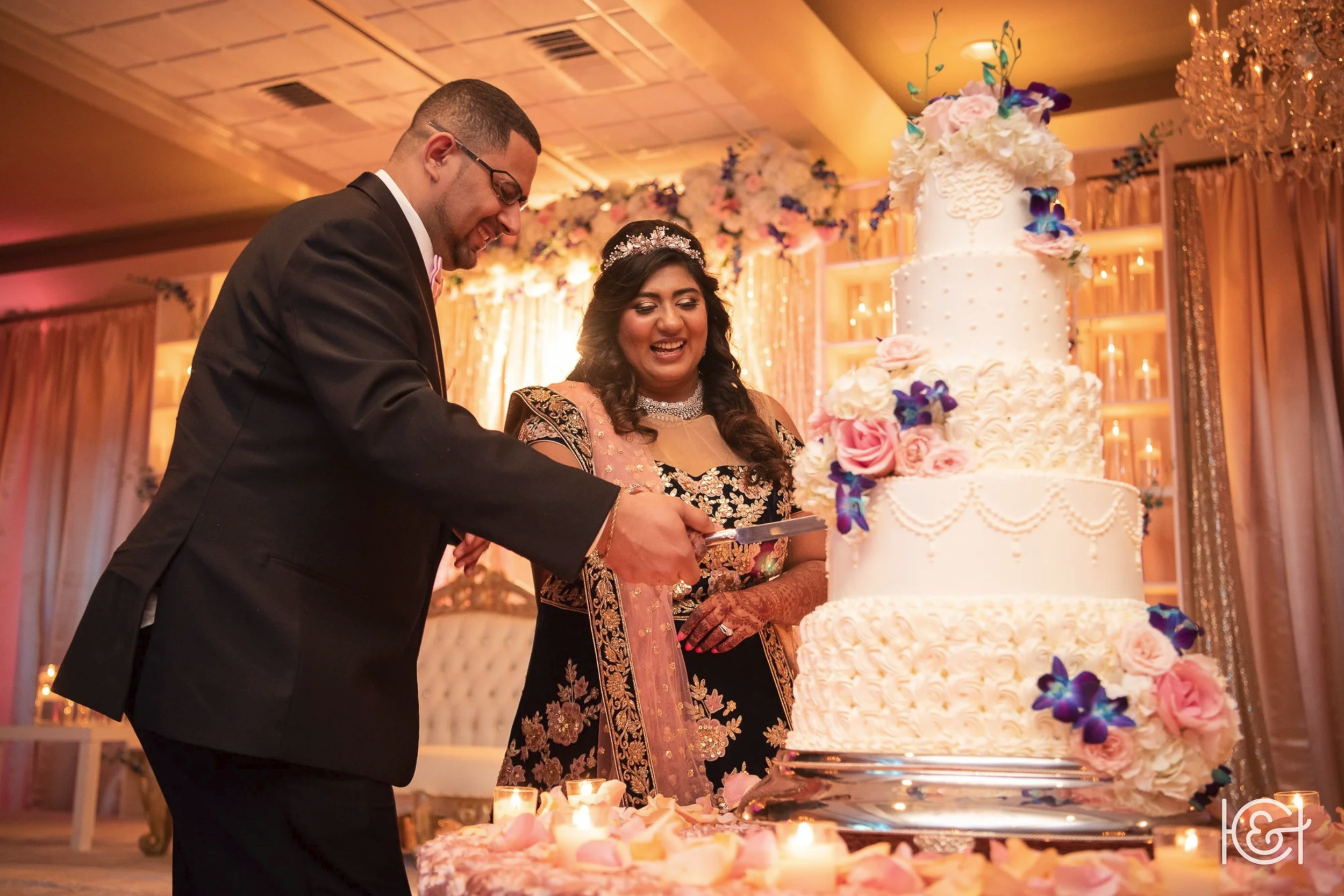 A couple cutting a wedding cake with a decorated four-tier cake decorated with pink roses and purple flowers, in a warmly lit reception hall.
