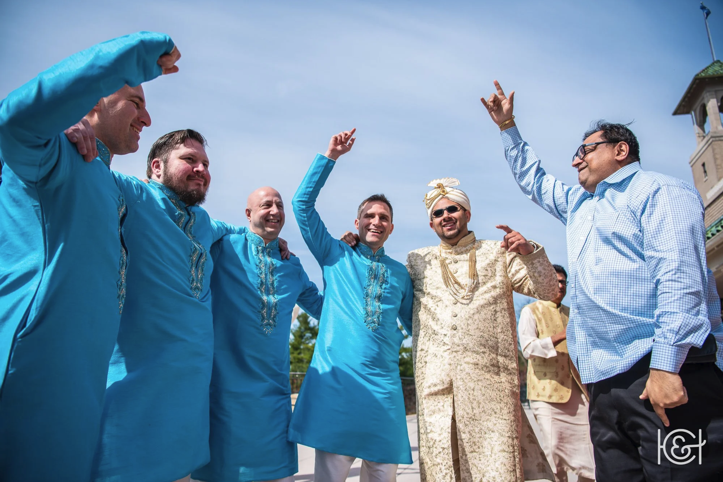 Group of men celebrating outdoors, some dressed in traditional South Asian attire and others in casual shirts, with arms raised in joy under a partly cloudy sky.