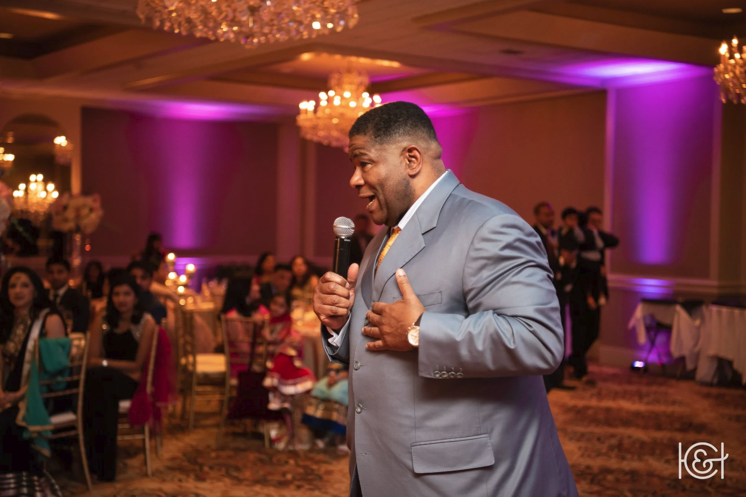 A man giving a speech or toast at a formal event or celebration, holding a microphone with one hand and resting the other hand on his chest, in a decorated banquet hall with purple lighting and chandeliers, surrounded by seated guests.