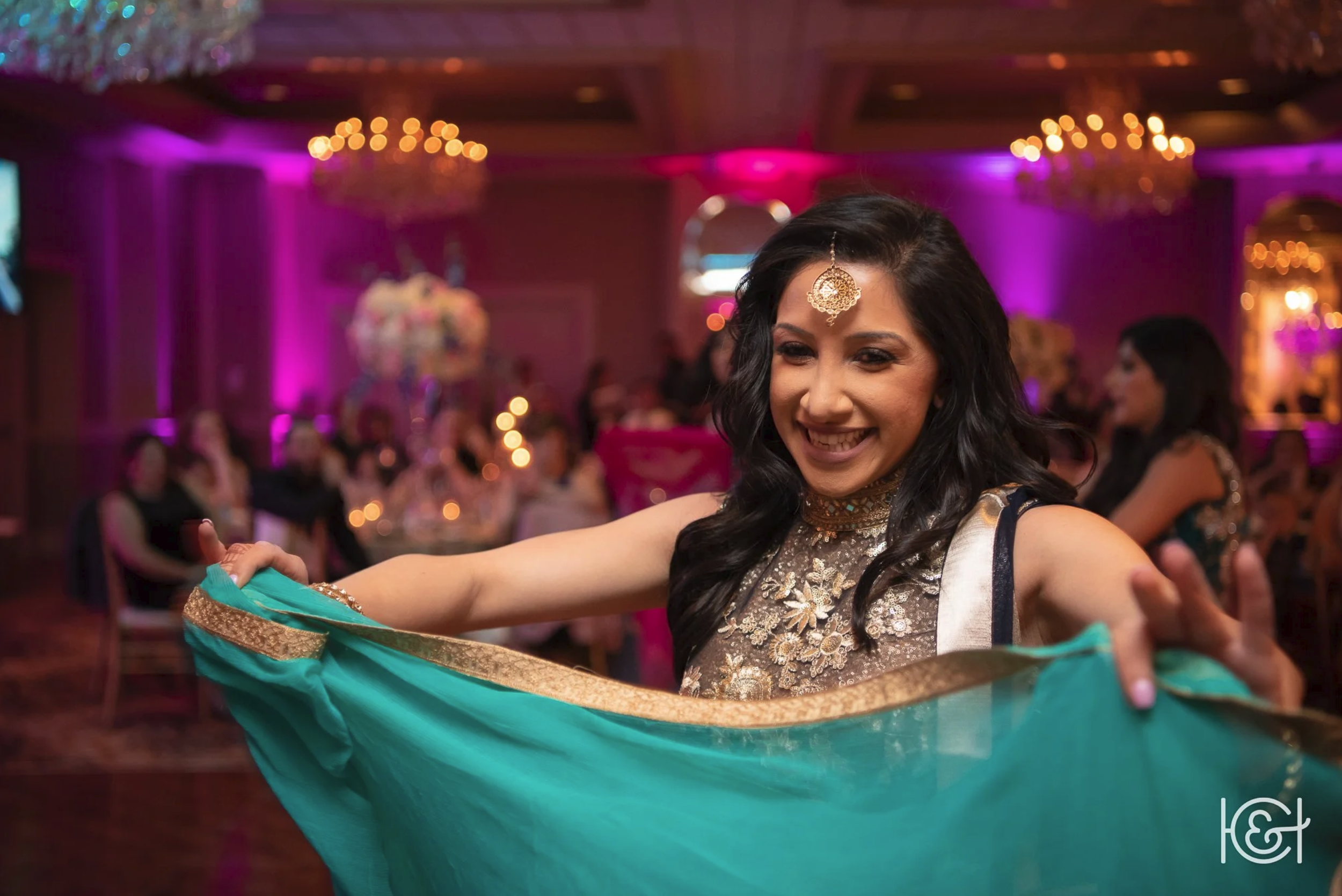 Woman in a traditional Indian outfit dancing with a turquoise dupatta at a decorated wedding reception with pink and purple lighting.