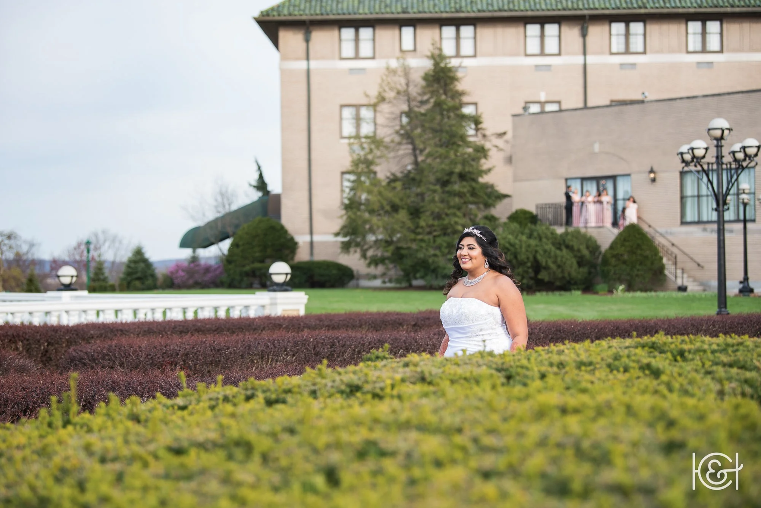 A bride in a white wedding dress with jewelry, smiling and standing outdoors on a lawn with bushes and trees, with a large building, lamp posts, and guests in the background.