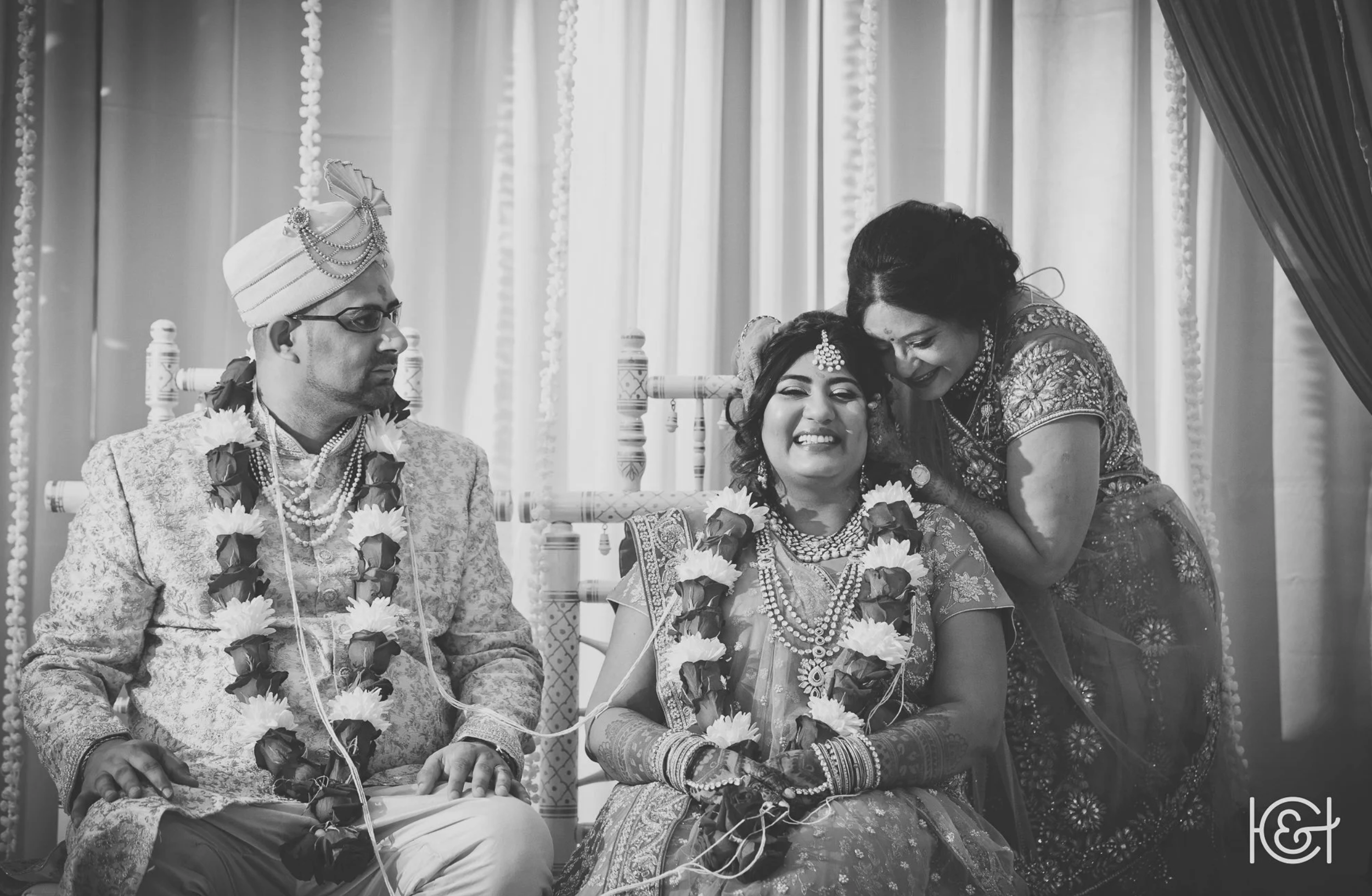 Black and white photograph of a joyful Indian wedding scene. The bride is seated, smiling with joy, adorned with traditional jewelry including a maang tikka and bangles, and wearing a floral garland. The groom sits beside her, looking at her, wearing