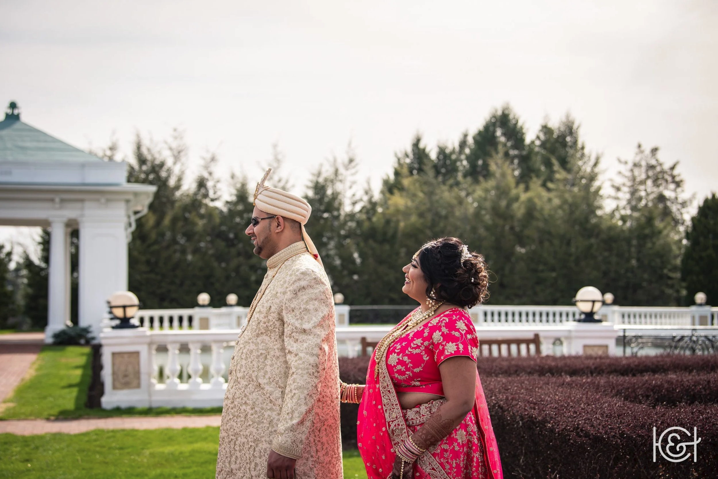 A couple dressed in traditional Indian wedding attire standing outdoors in front of a white pavilion, with the man leading and the woman smiling behind him.