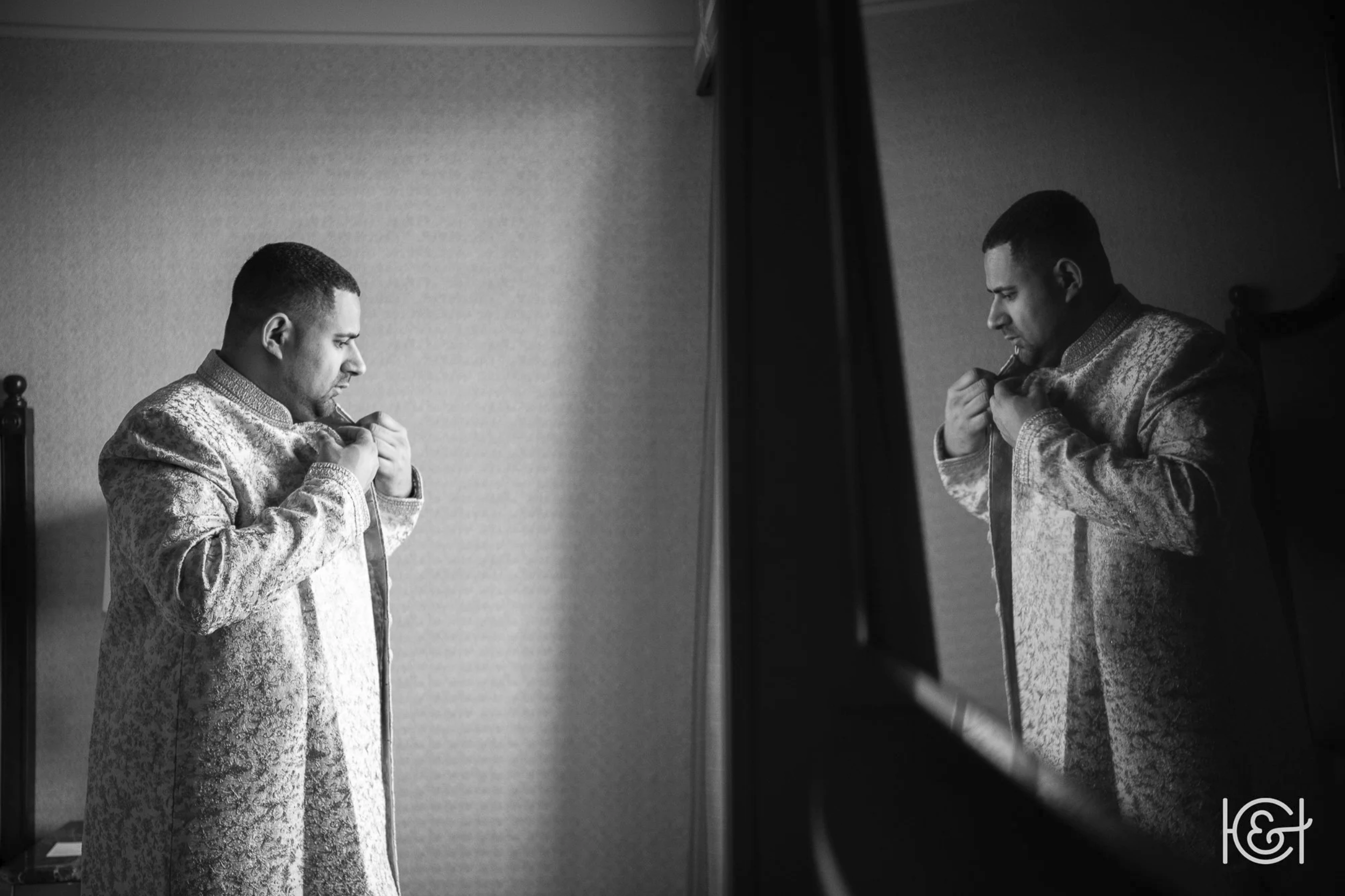 A man wearing a textured long coat is standing in front of a mirror, adjusting his collar.