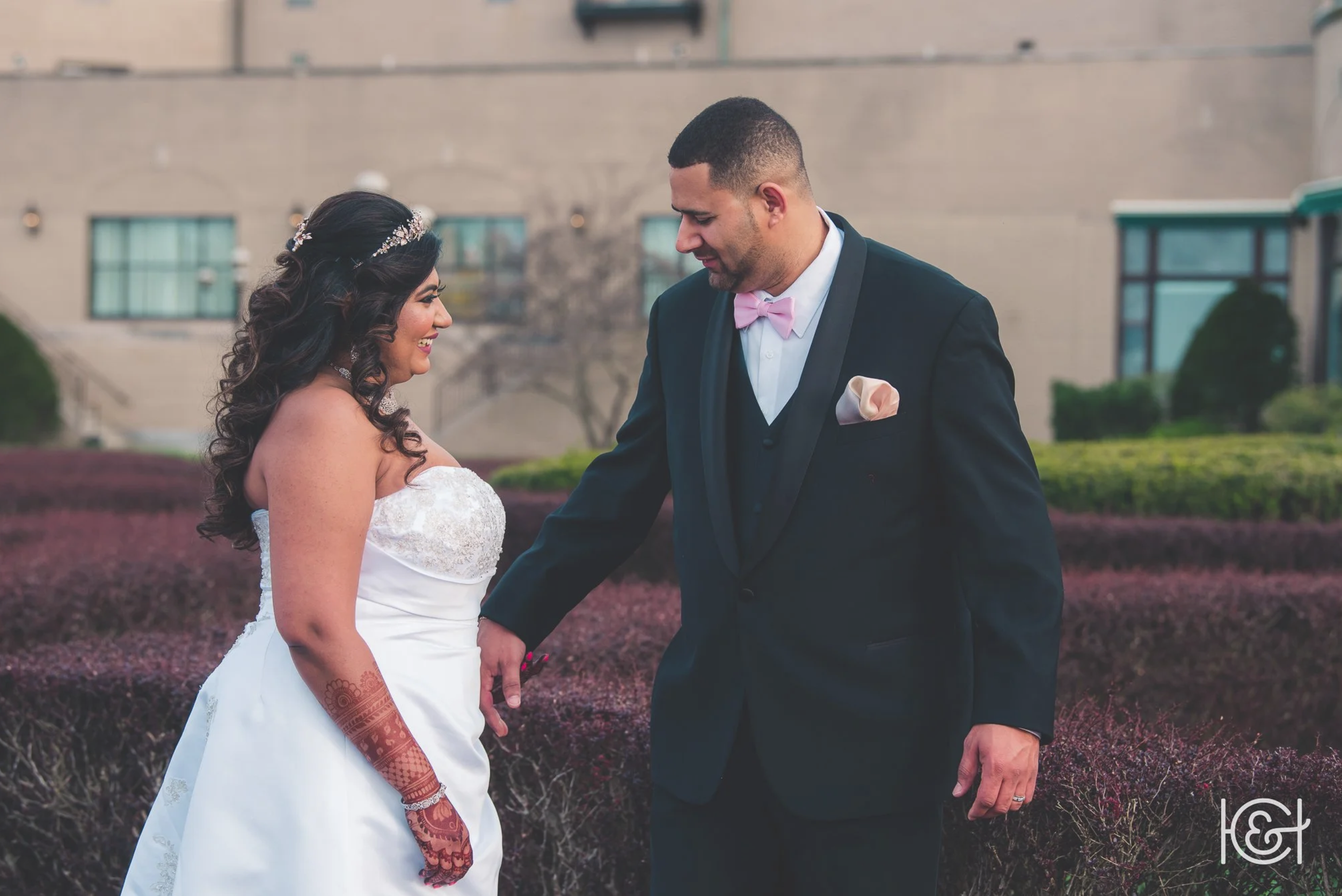 Bride and groom holding hands outdoors, wedding attire, smiling, with a building and bushes in the background.