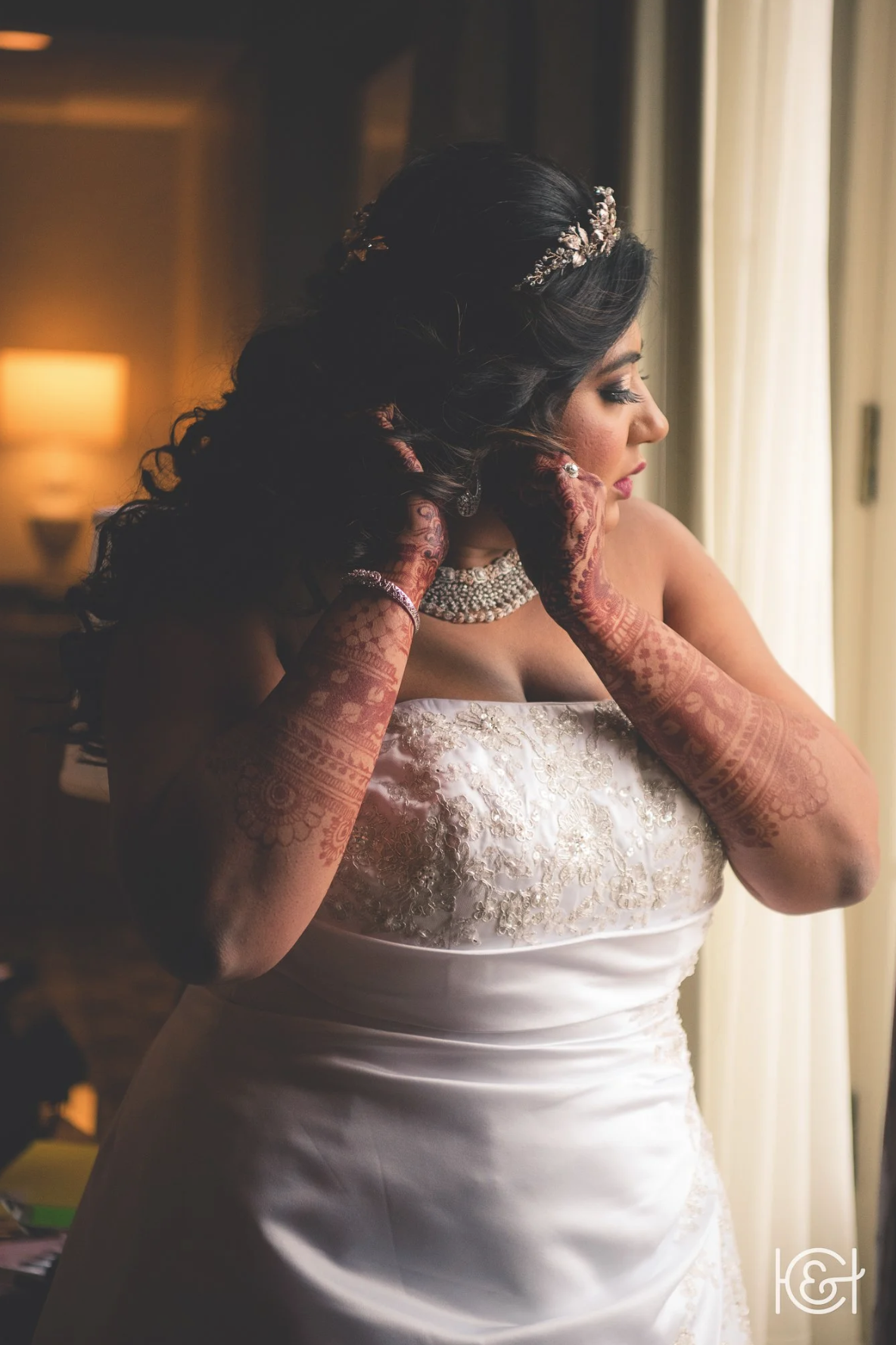 A bride adjusting her earring near a window, wearing a strapless wedding gown and intricate henna designs on her arms.