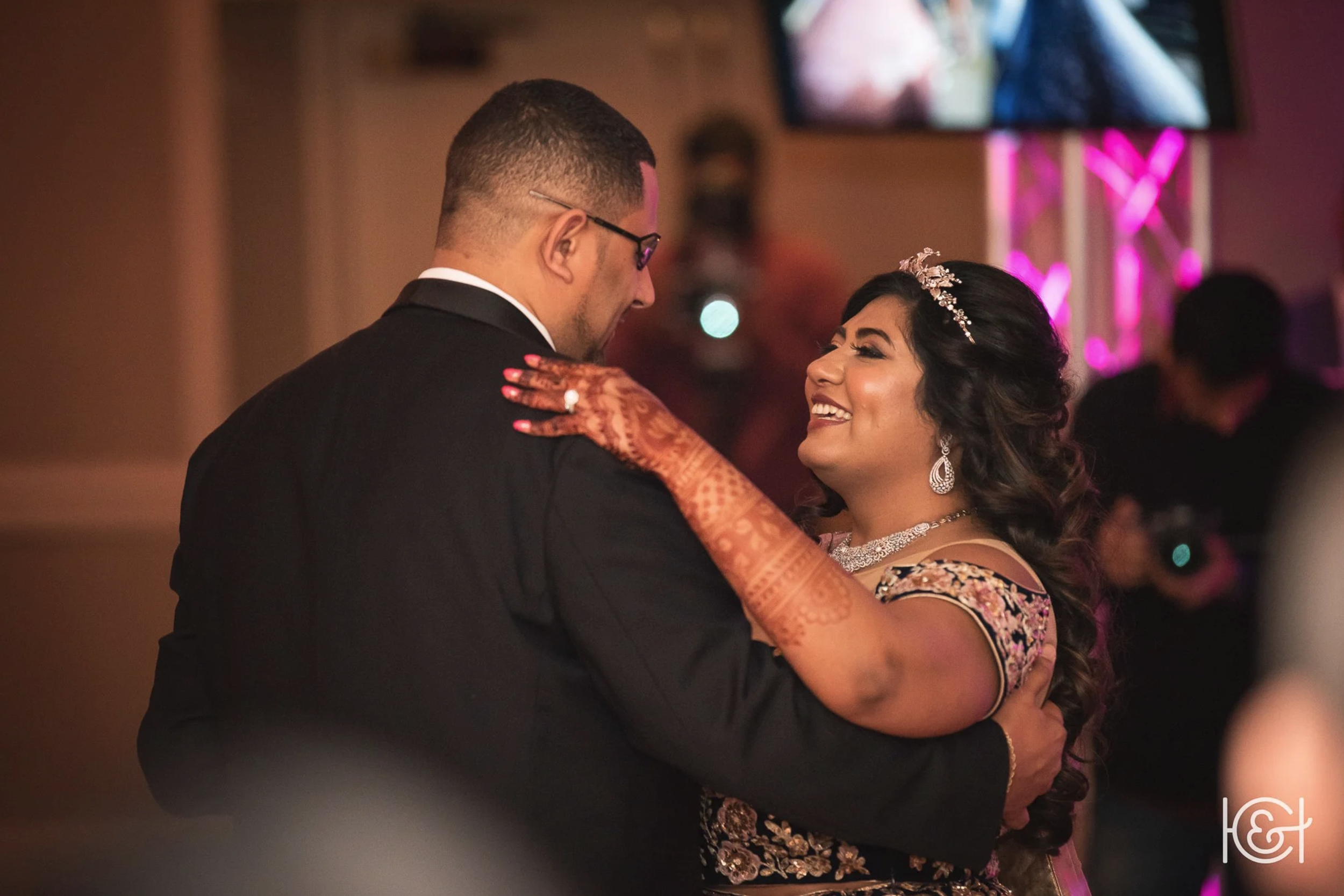 A couple dancing at a wedding reception, with the woman wearing traditional South Asian attire and jewelry, and the man dressed in a tuxedo.