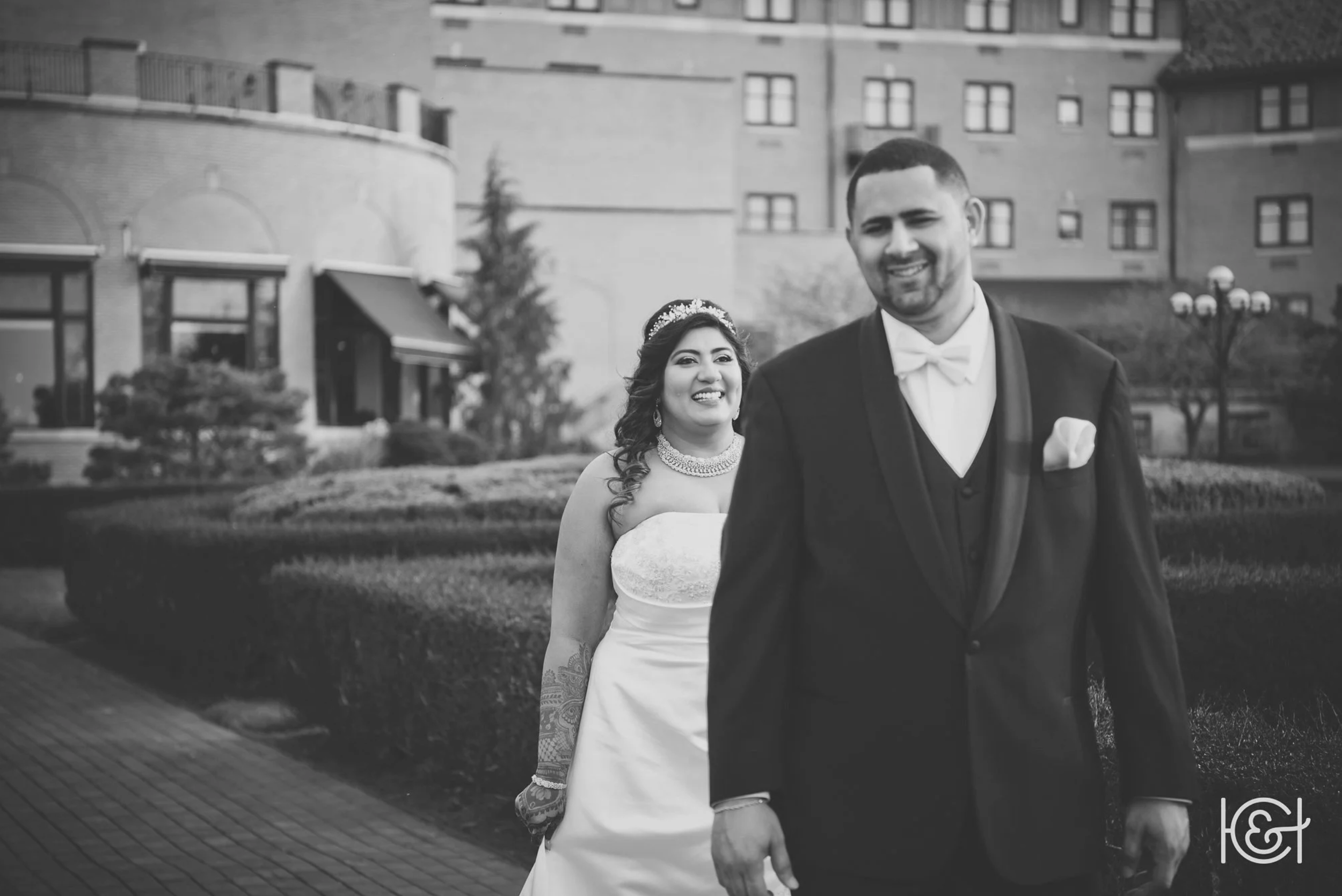 A black and white photo of a smiling bride in a strapless wedding dress and a groom in a tuxedo, standing outdoors with a city building in the background.
