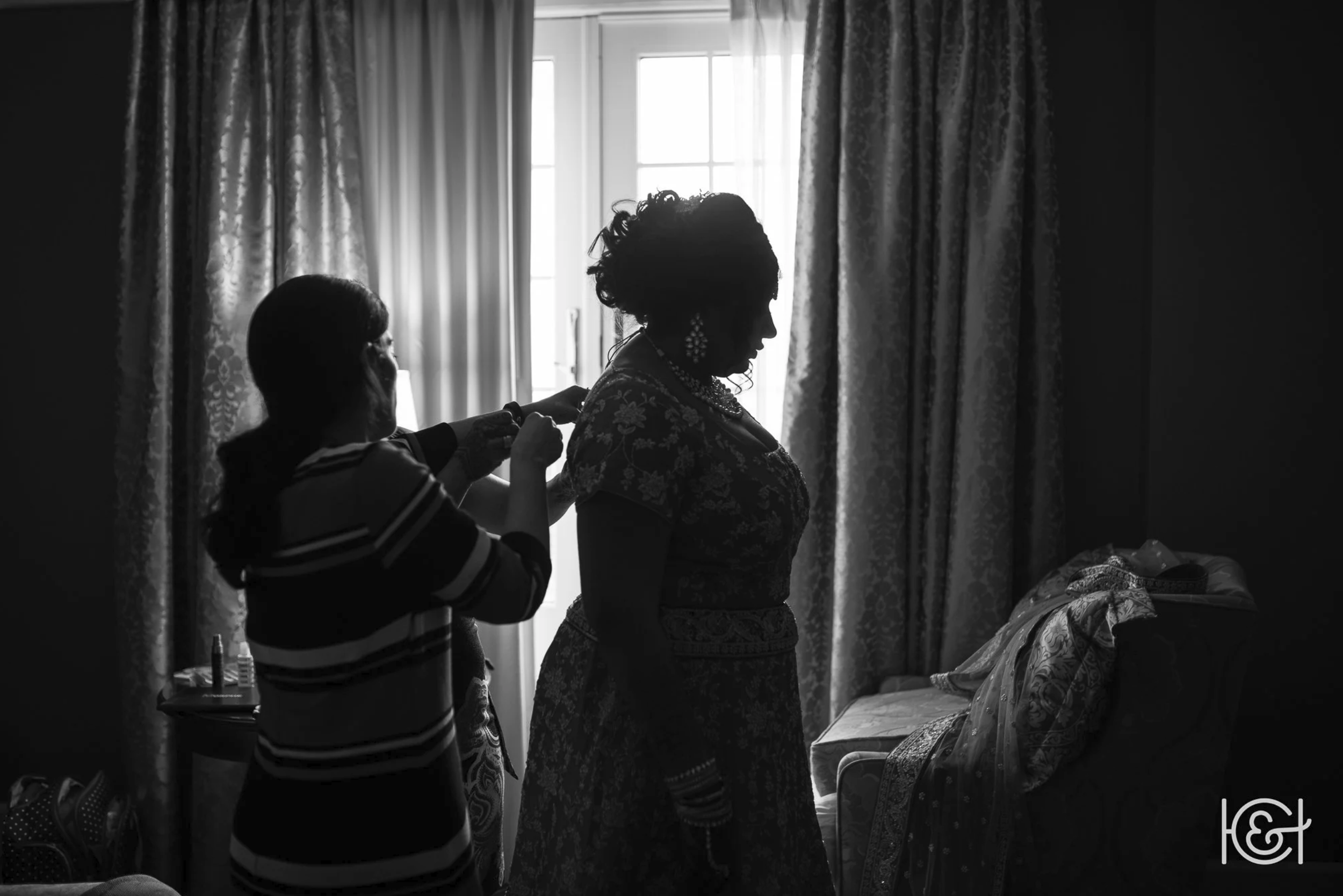 Silhouette of a woman being helped with her jewelry, with another person assisting, in a room with curtains and a window in the background.