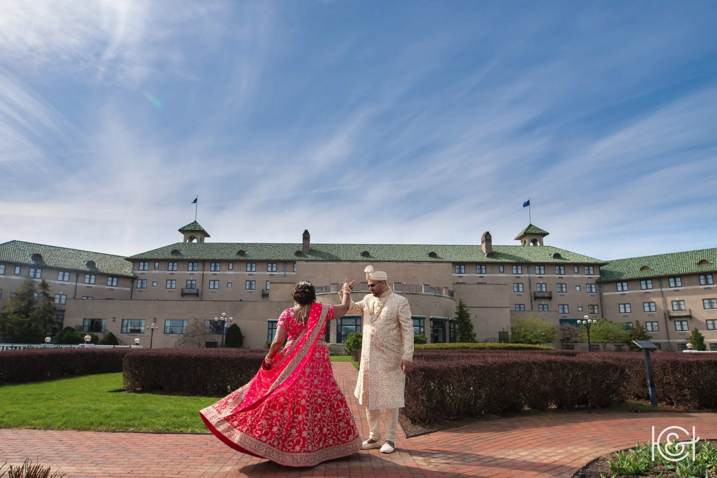 A couple dressed in traditional Indian wedding attire dancing outdoors in front of a large hotel or building with a green roof and blue flags, on a sunny day with a clear blue sky.
