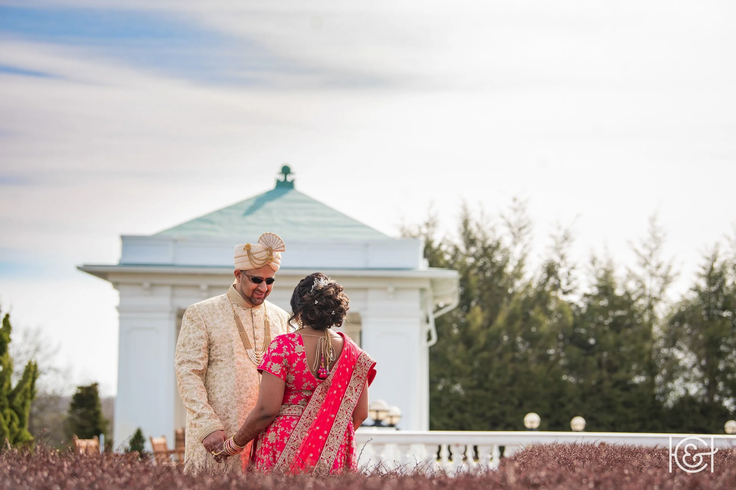 A couple dressed in traditional wedding attire holding hands outdoors in front of a white gazebo, with trees and a partly cloudy sky in the background.