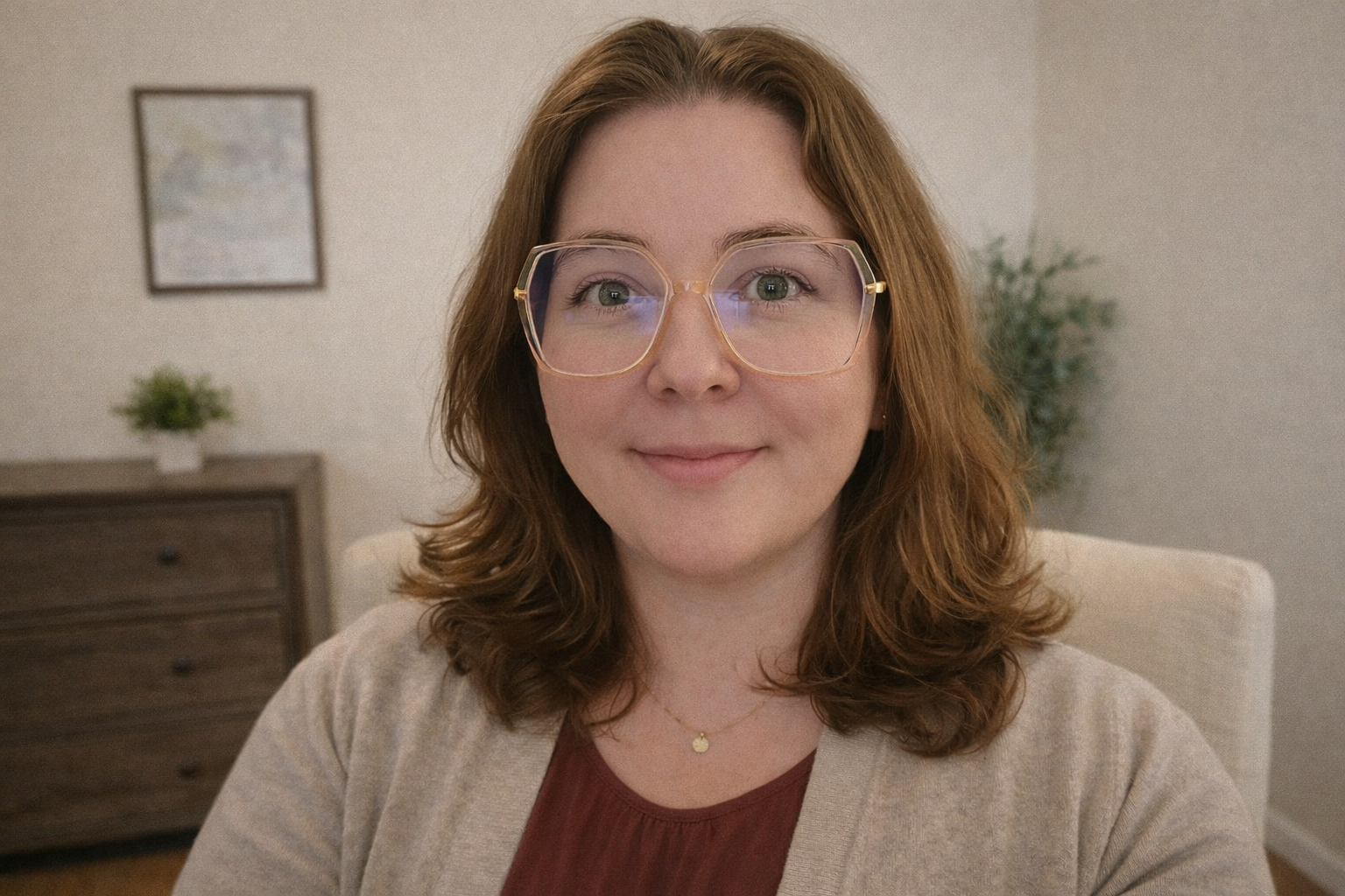 A woman with shoulder-length wavy auburn hair and glasses with clear frames, smiling softly at the camera, in a cozy indoor setting with a beige wall, a picture frame, a wooden dresser, and a potted plant in the background.