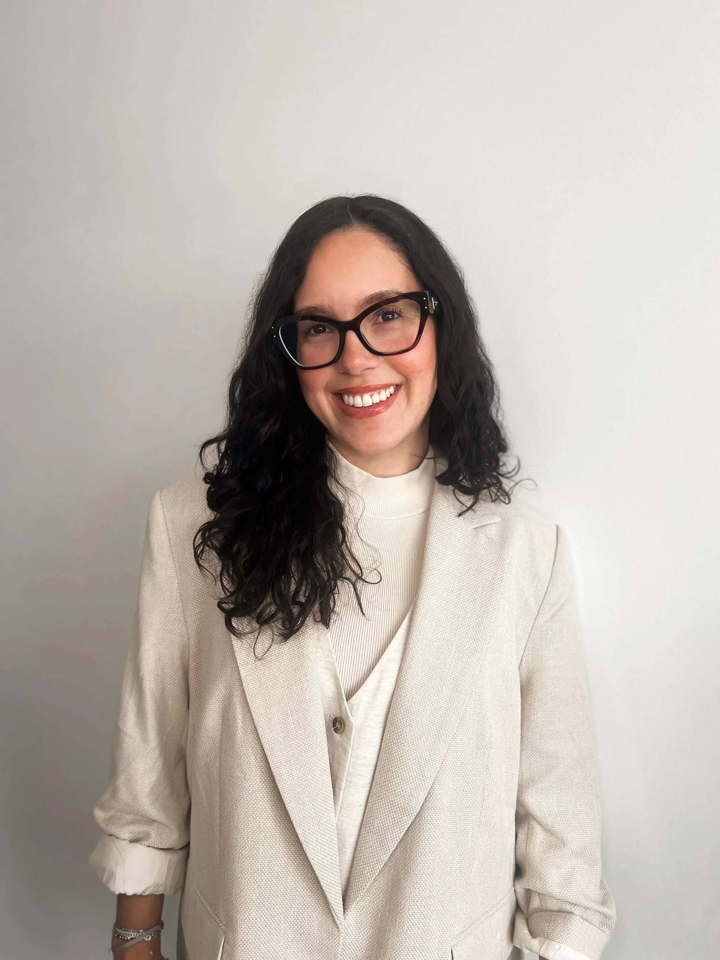 A woman with dark curly hair, wearing glasses and a white blazer, smiling in front of a plain white wall. Dr. Sasha Shifrin.