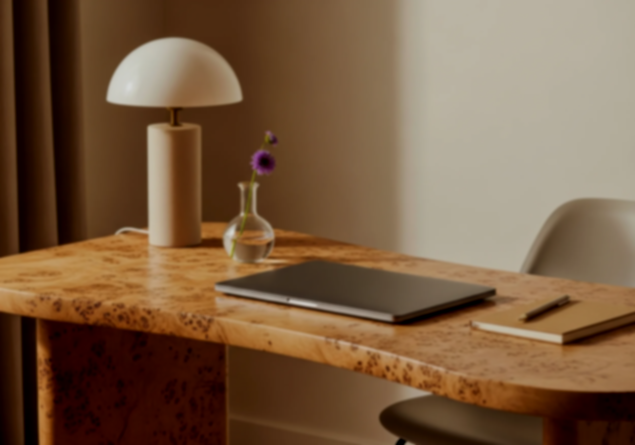 A wooden desk with a modern white table lamp, a small vase with purple flowers, a closed laptop, a notebook, and a pen, next to a beige chair, in a softly lit room.