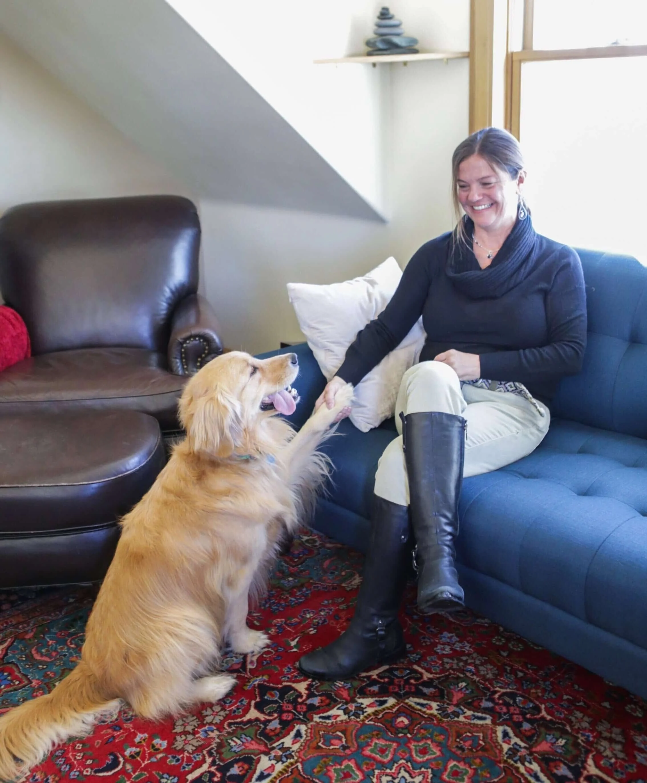 A woman sitting on a blue couch smiling as she holds hands with a golden retriever dog, which is sitting on a red patterned rug, in a cozy living room with a brown leather chair and decorative shelves.