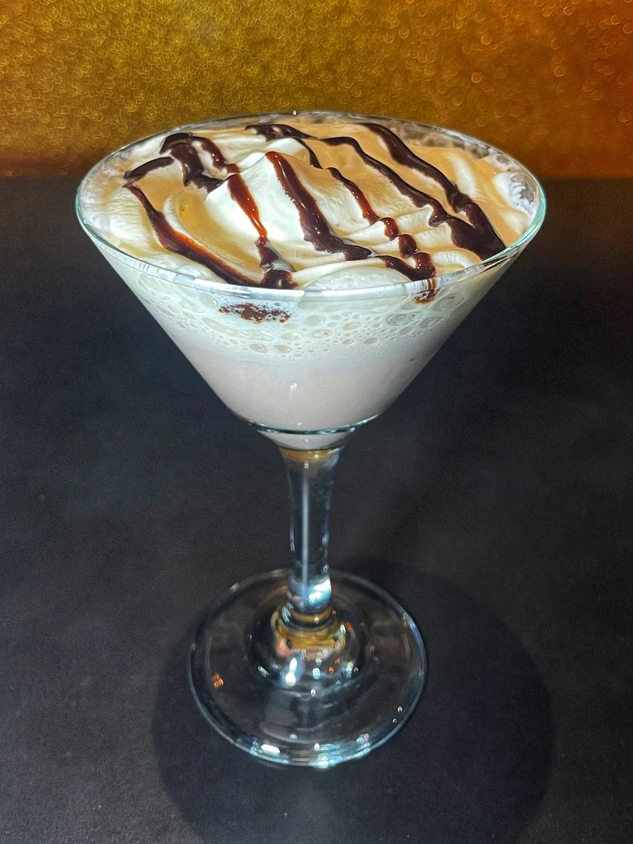 A creamy, frothy cocktail in a martini glass topped with whipped cream and drizzled with chocolate syrup.