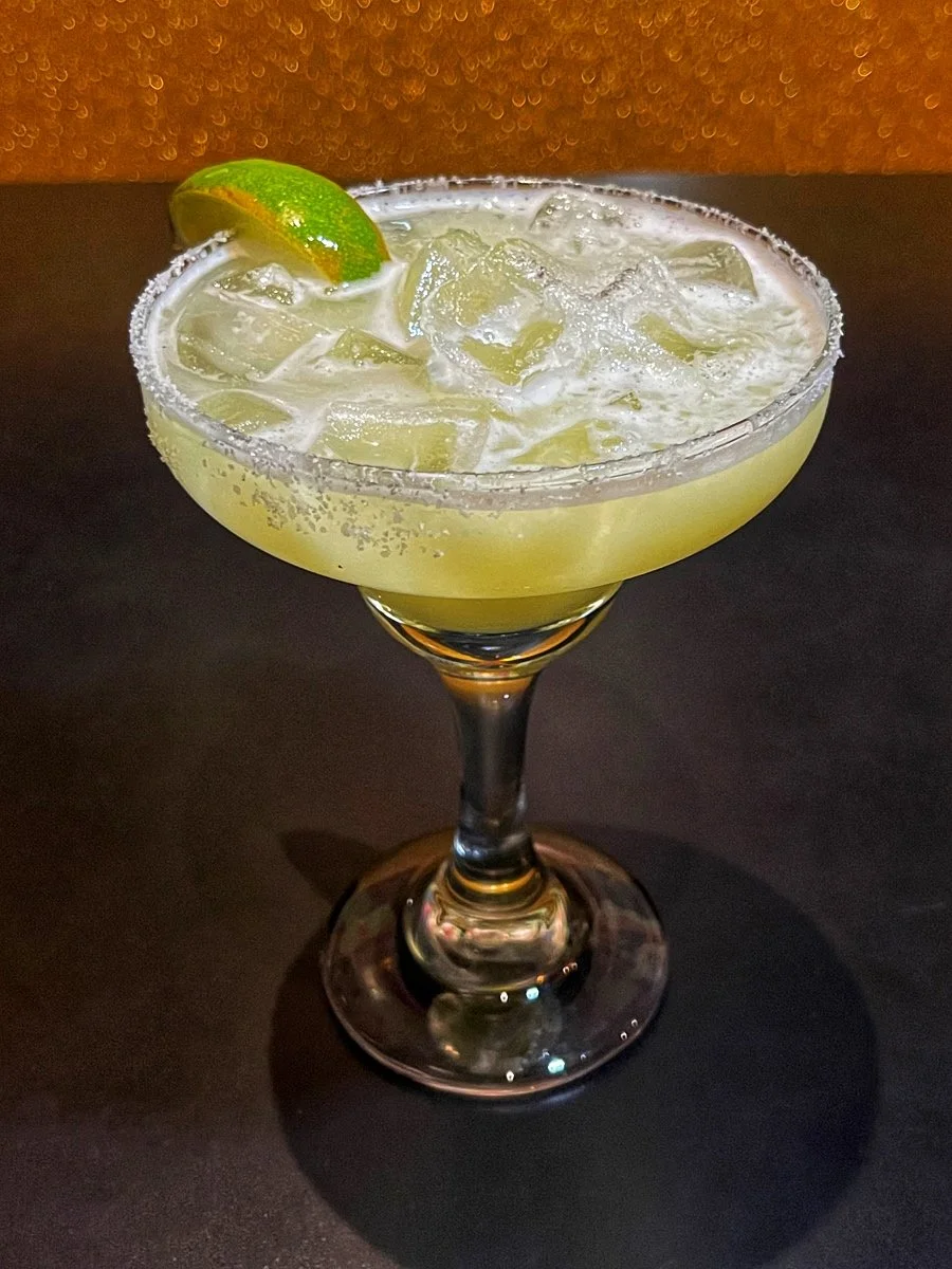 A cocktail glass with salted rim, filled with a light yellow drink and ice, garnished with a lime wedge.