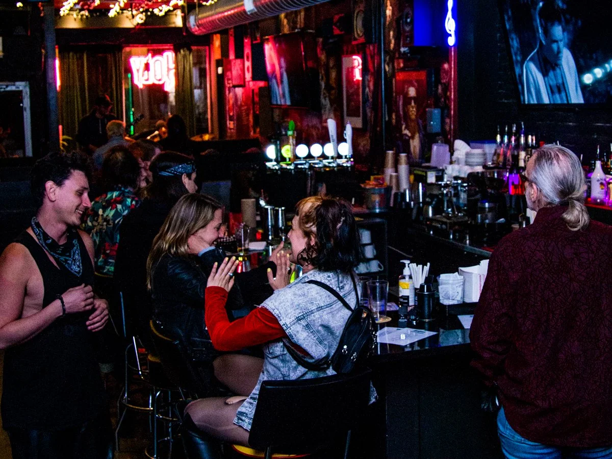 People enjoying drinks and conversing at a bar with colorful neon signs and television screens in the background.