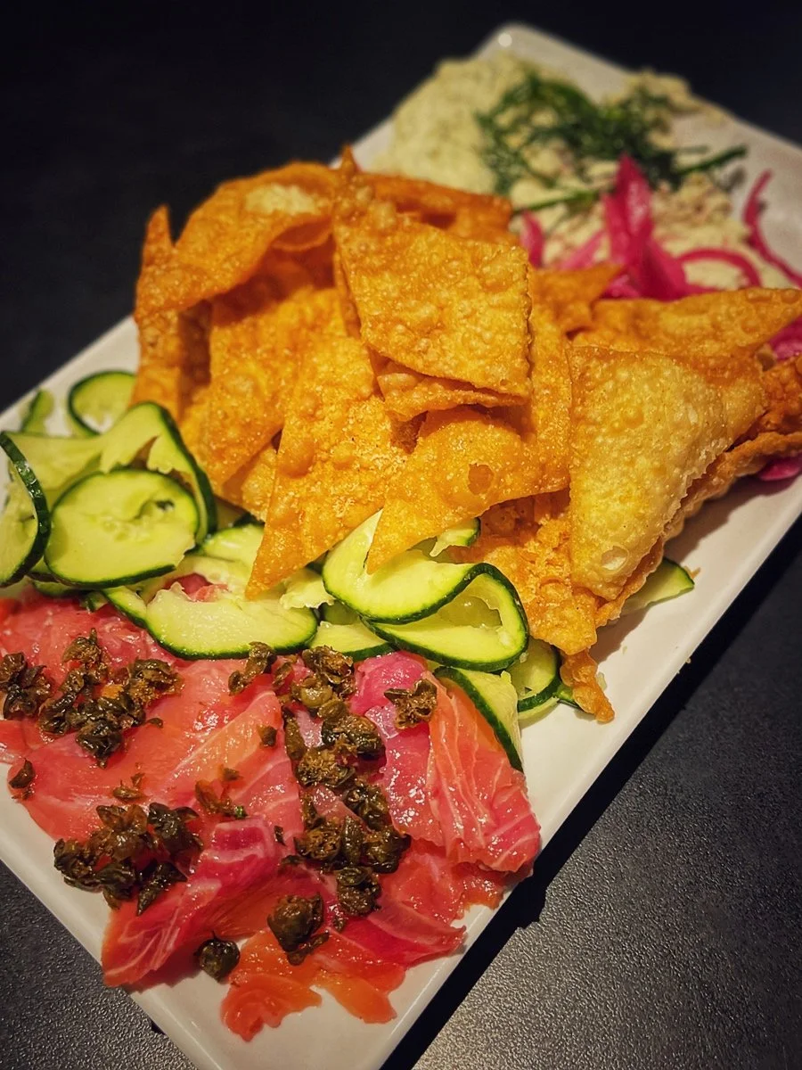 A rectangular plate with nachos, cucumbers, smoked salmon with capers, and green dip garnished with herbs.