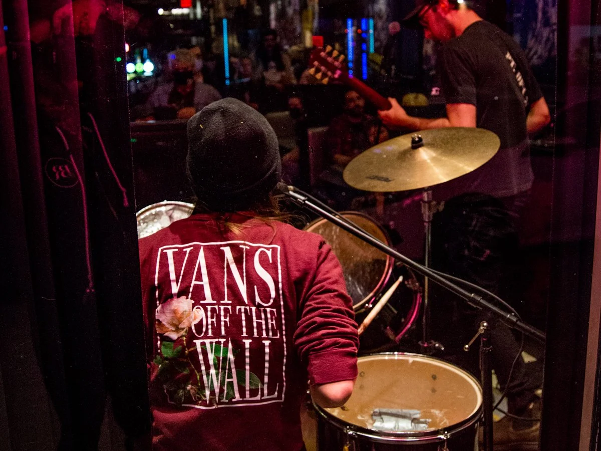 A band performing live in a dimly lit venue, viewed from behind. The audience is visible through the glass window, enjoying the performance.