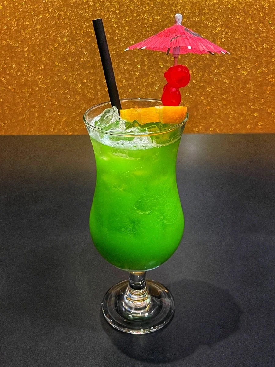 Green tropical cocktail in a tall glass garnished with a slice of orange and maraschino cherries, with a paper umbrella and black straw, on a dark surface with a gold, textured background.