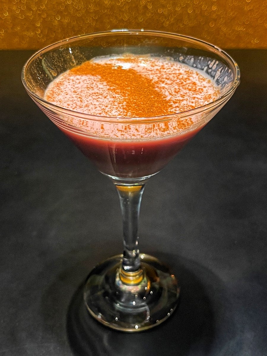 A cocktail in a martini glass with a red liquid, topped with a sprinkle of ground spice, possibly cinnamon or nutmeg.