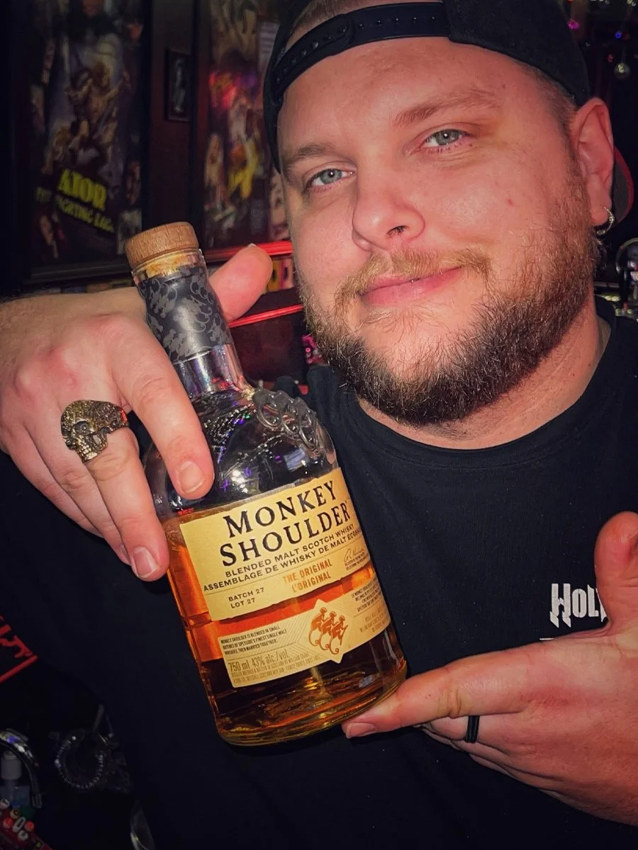 A man with a beard and baseball cap holding a bottle of Monkey Shoulder whisky in a bar.