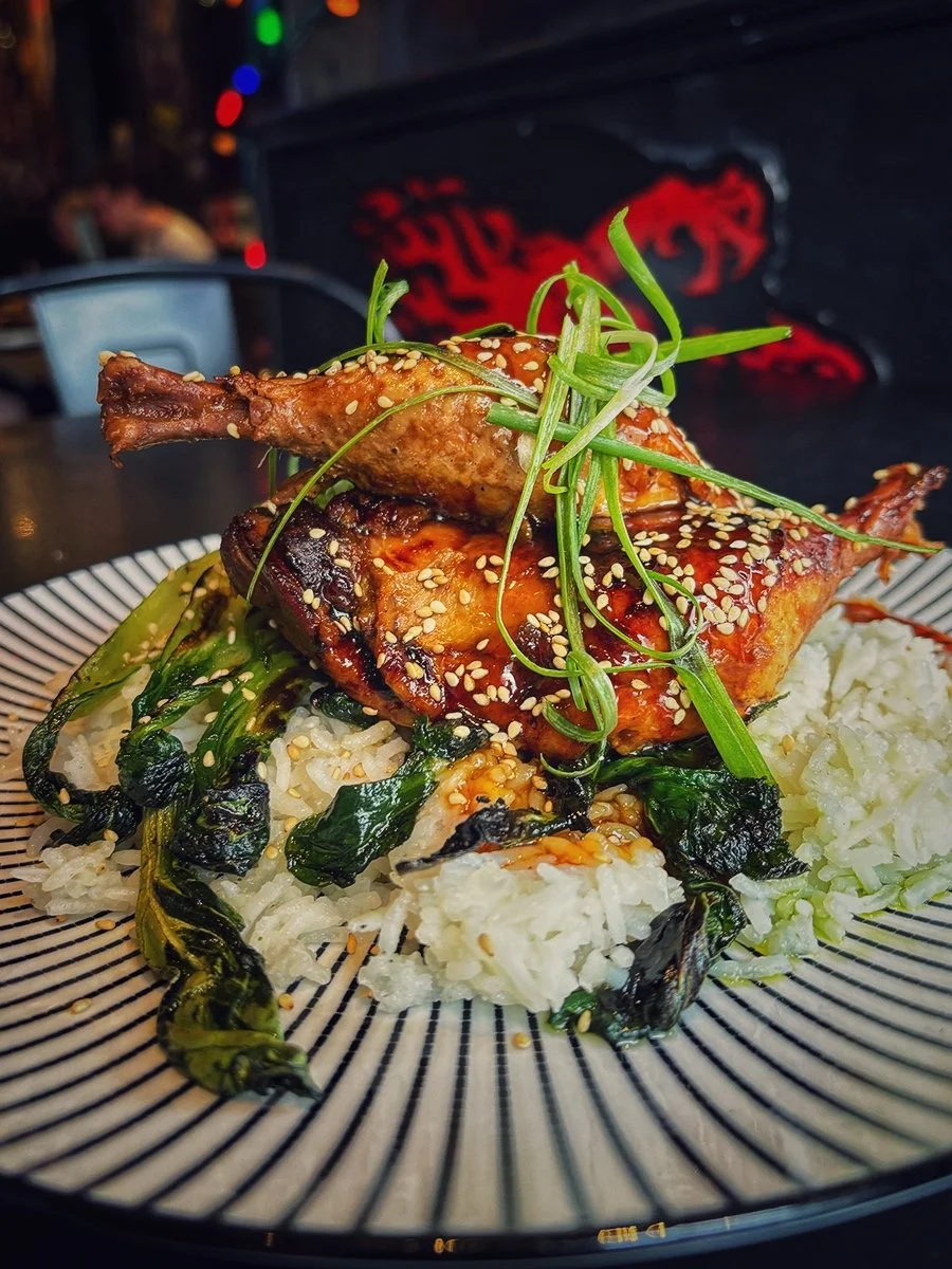 A plate with grilled chicken topped with green onions and sesame seeds, served over white rice and sautéed greens, on a striped black-and-white plate.