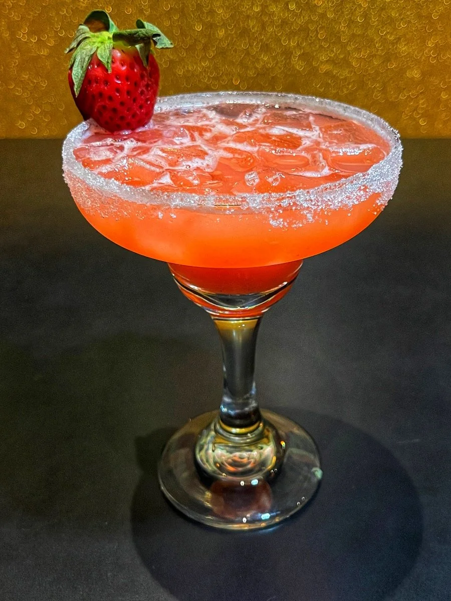 Pink cocktail with ice in a glass, rimmed with sugar, garnished with a strawberry, on a dark surface with a gold textured background.