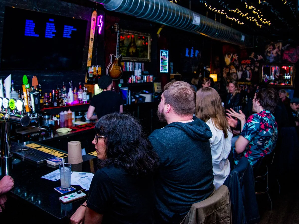 People sitting at a bar counter in a dimly-lit music-themed bar or club, with bottles and drinks on the counter, a guitar hanging on the wall, and neon signs.