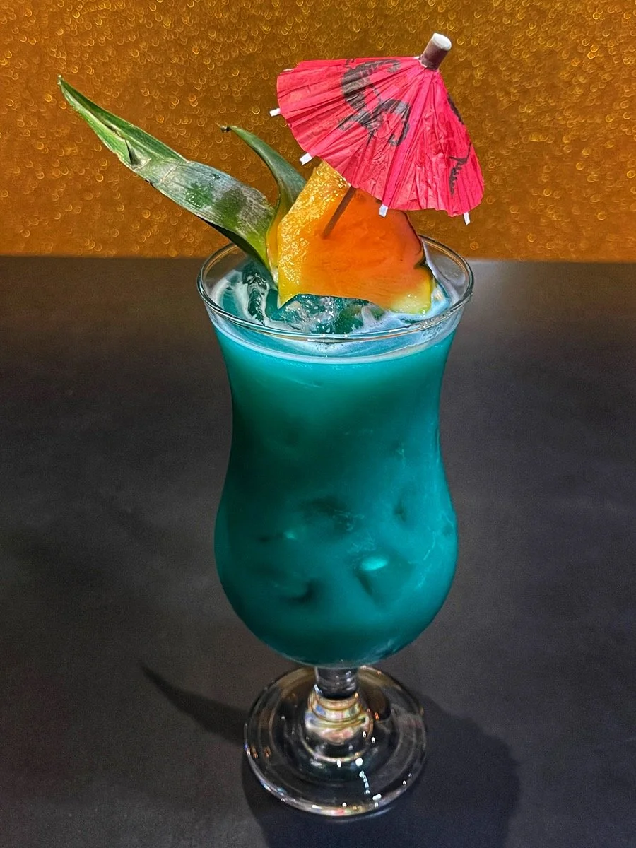 A blue tropical cocktail in a tall glass. It is garnished with a fruit slice, a pineapple leaf, and a small red paper umbrella.