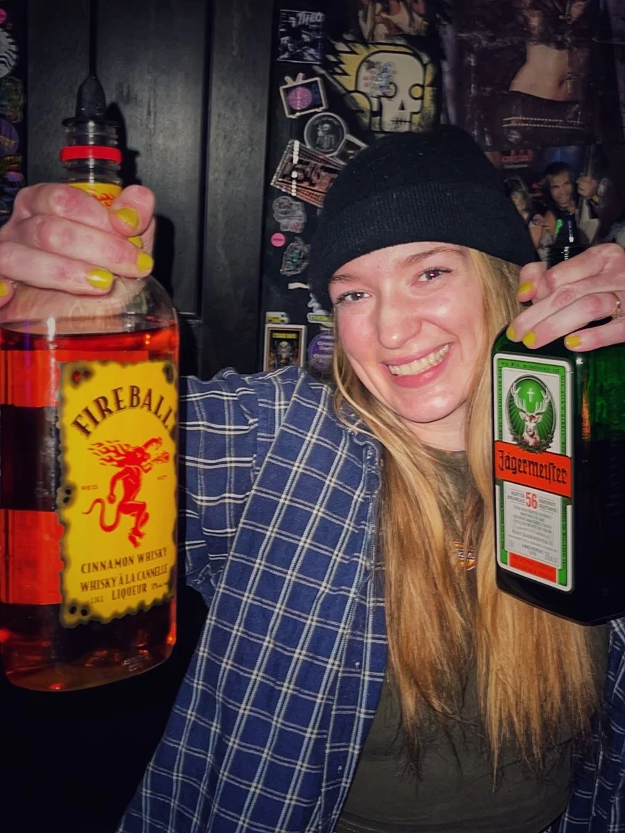 A smiling woman with long blonde hair wearing a black beanie and a plaid shirt, holding a bottle of Fireball cinnamon whisky in one hand and a bottle of Jägermeister in the other, in a bar setting.