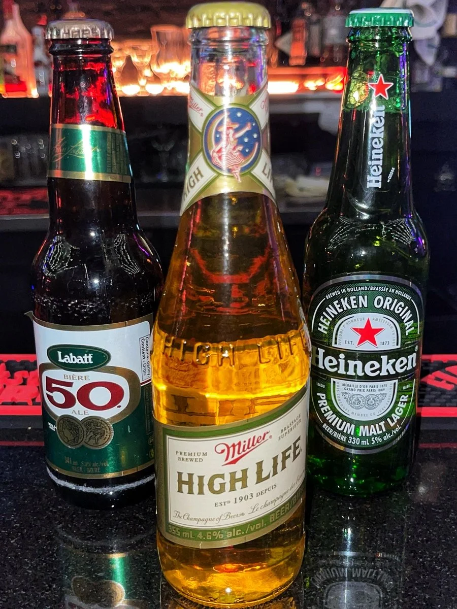 Three beer bottles on a bar counter, with the middle one labeled 'Miller High Life,' and flanked by a dark bottle on the left labeled 'Labatt 50' and a green Heineken bottle on the right.