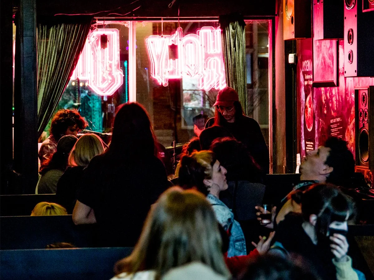 Crowd of people inside a dimly lit bar or restaurant with a neon sign reading 
