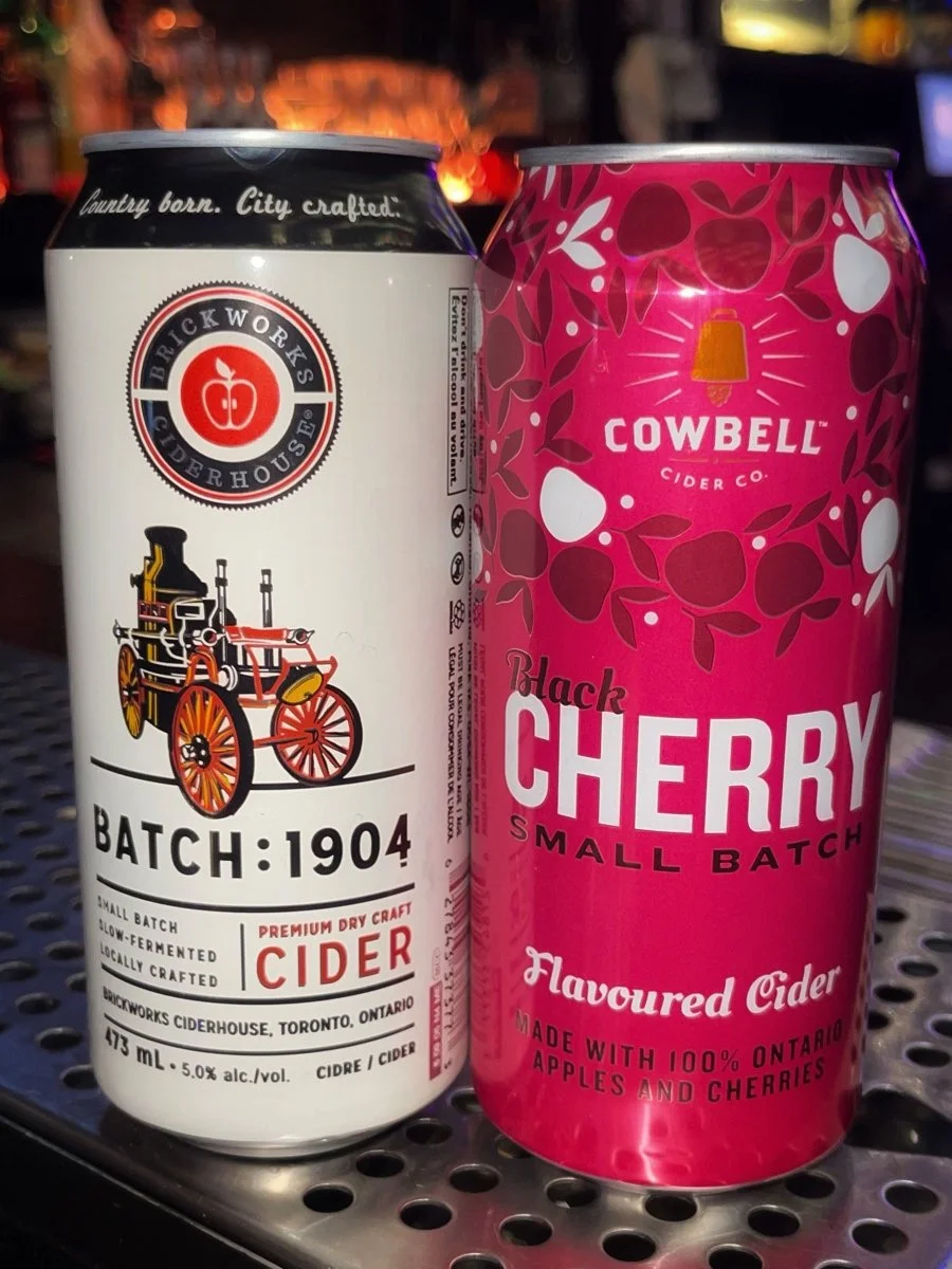 Two cans of cider, one from Brickworks Ciderhouse with a vintage wagon logo and one from Cowbell Cider Co. with a bright pink label and cherry design.