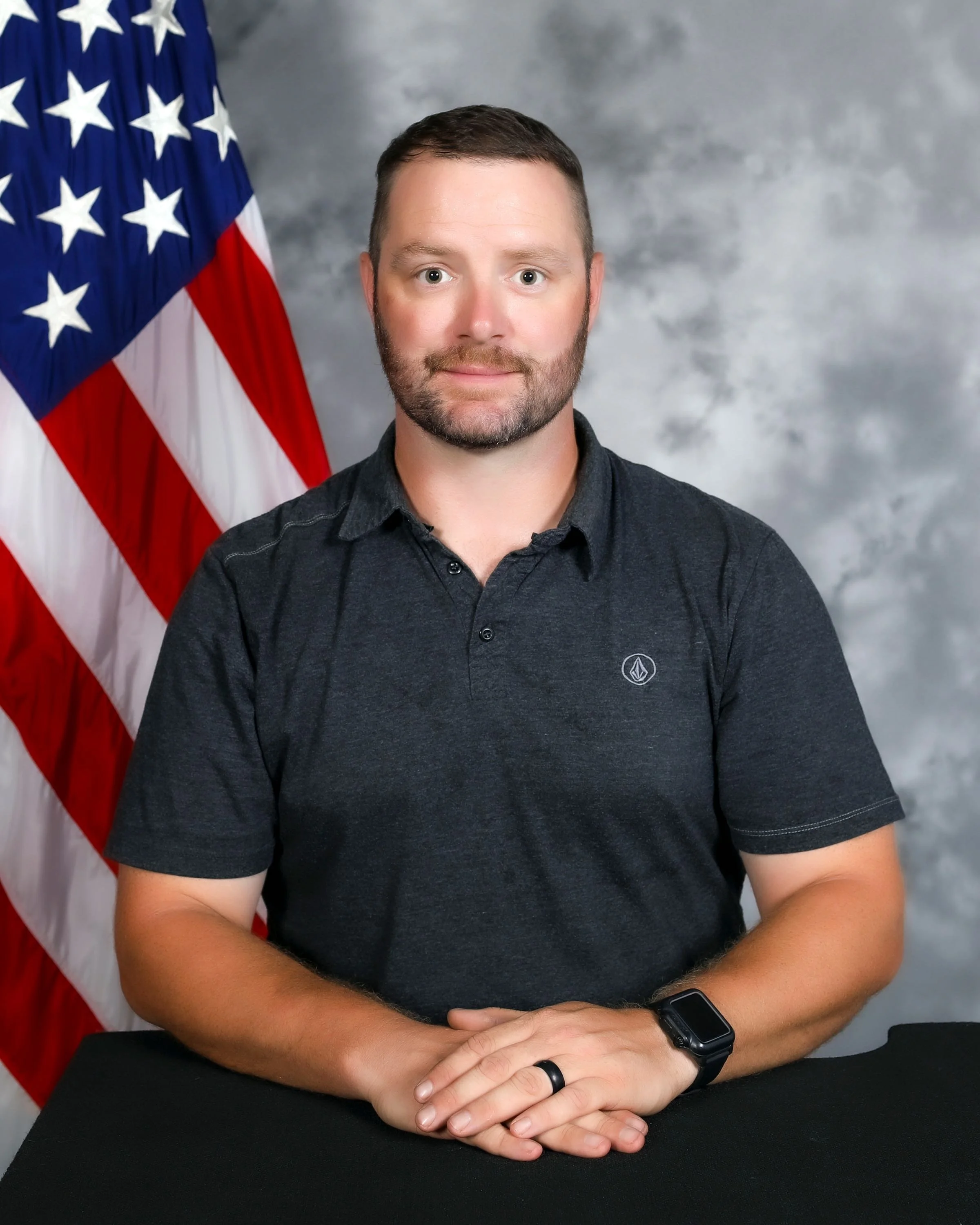 A man with short brown hair and a beard, wearing a dark gray polo shirt and a smartwatch, seated with clasped hands, in front of an American flag and a gray mottled background.
