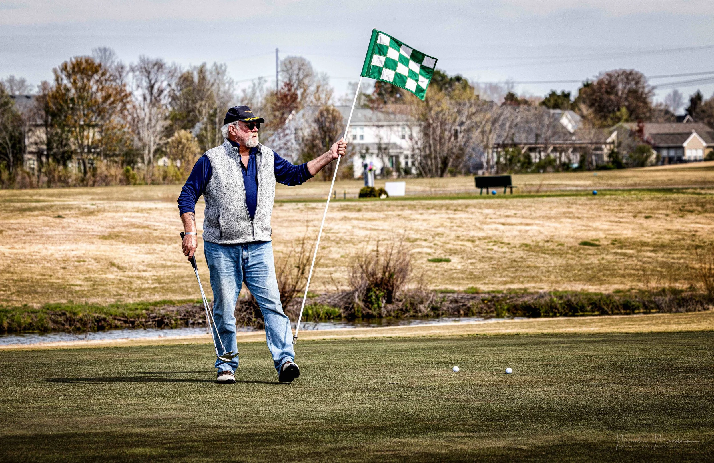 An older man playing golf on a golf course, holding a checkered flag, with houses and trees in the background.