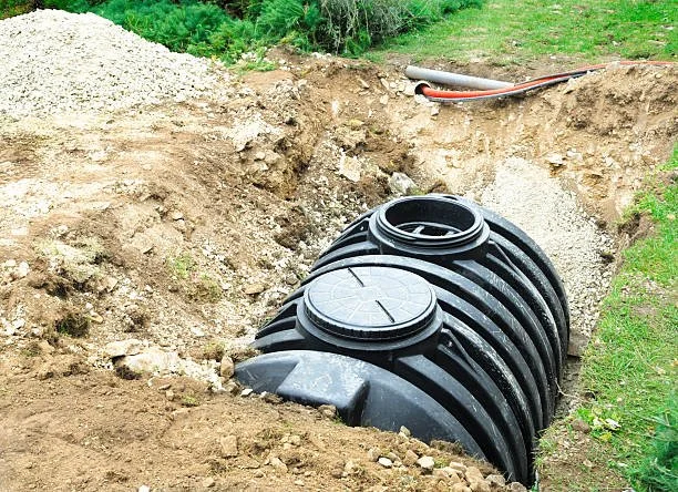 Underground black plastic septic tank installed in a dug trench with surrounding dirt and some tubing nearby.