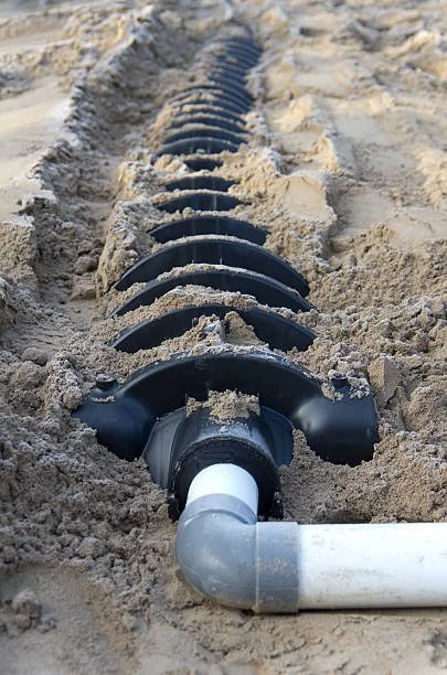 A close-up of a pipe installation in the sand with a black plastic pipe and a joint, surrounded by a trench in the sand.