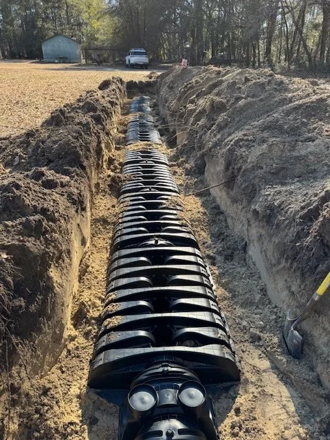 A sewer pipe being installed in a trench outdoors, with trees, a building, and a car in the background.