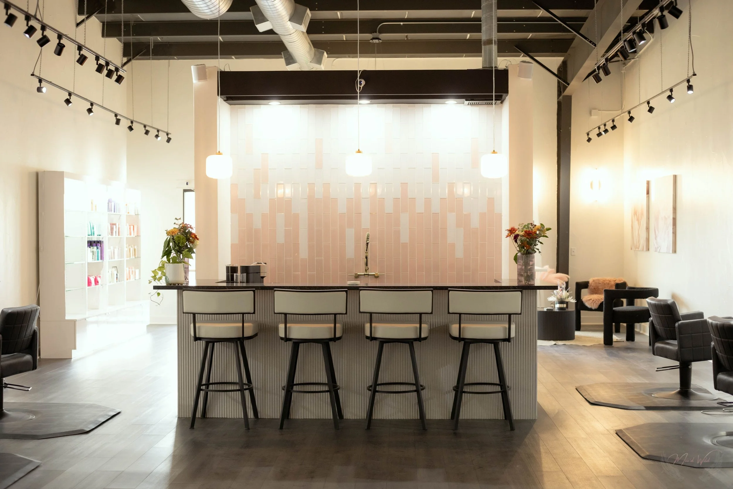 Modern salon interior with a central bar area, four bar stools, pink tiled wall, hanging lights, flower vases, shelves with products on the left, and styling chairs on the right.